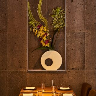 Restaurant dining table set for six with plates, napkins, and glasses, and a framed vertical floral arrangement on a dark stone wall behind.