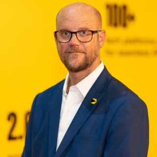 Portrait of a bald man with glasses in a blue suit and white shirt, standing against a bright yellow background.
