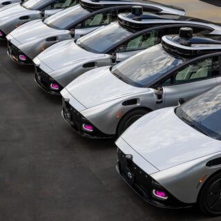 Row of silver Lyft self-driving cars with rooftop sensors parked in a lot, aligned in a line.