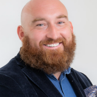 Portrait of a bald man with a thick red beard, smiling, wearing a blue shirt and navy blazer against a light background.