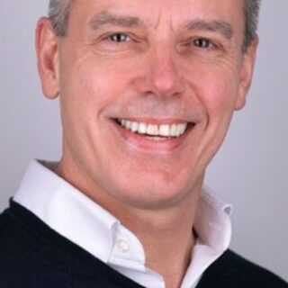 Smiling man in a white collared shirt and dark sweater, head-and-shoulders portrait against a gray background.