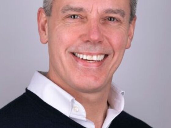 Smiling man in a white collared shirt and dark sweater, head-and-shoulders portrait against a gray background.