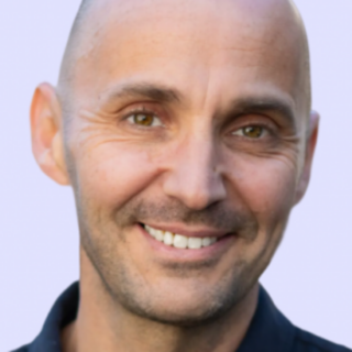 Close-up portrait of a smiling man with a shaved head against a pale purple background.