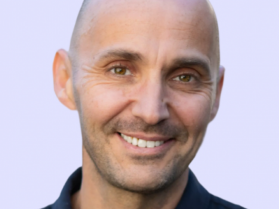 Close-up portrait of a smiling man with a shaved head against a pale purple background.