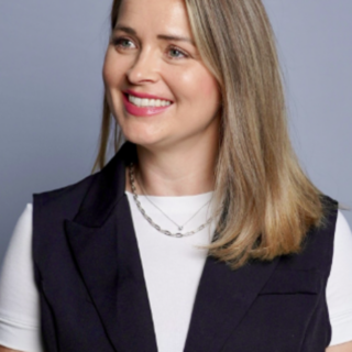 Smiling woman with shoulder-length blonde hair wearing a white top and dark vest against a blue-gray background.