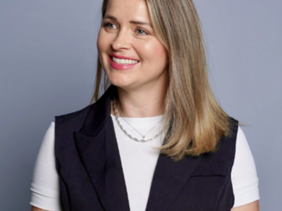 Smiling woman with shoulder-length blonde hair wearing a white top and dark vest against a blue-gray background.