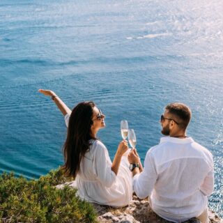 Couple sitting on a rocky cliff above the blue sea, toasting with champagne glasses.