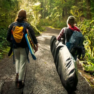 Two hikers walk a forest trail, each carrying gear and backpacks on a sunlit path surrounded by greenery and trees.