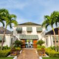 Entrance to a tropical resort villa with green-and-white striped awnings, palm trees, and a brick pathway leading to open doors.