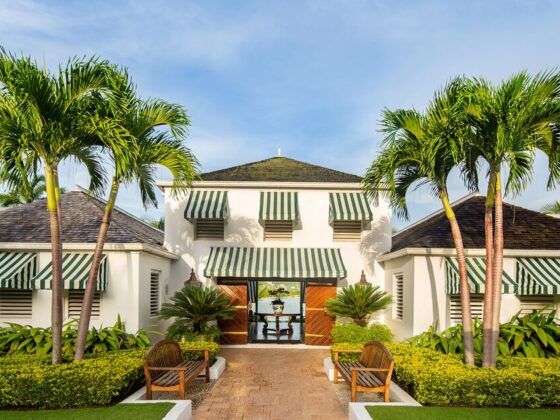 Entrance to a tropical resort villa with green-and-white striped awnings, palm trees, and a brick pathway leading to open doors.
