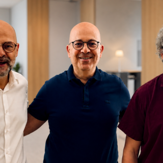 Three bald men with glasses smile at the camera, standing together in a bright, modern office lobby.