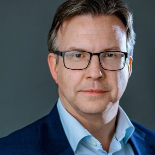 Professional headshot: man in a blue suit and light blue shirt wearing glasses, neutral dark background.