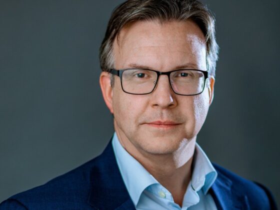 Professional headshot: man in a blue suit and light blue shirt wearing glasses, neutral dark background.