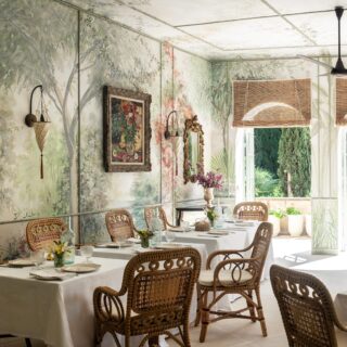 Sunlit dining area with white-clothed tables, wicker chairs, and a botanical mural wall.
