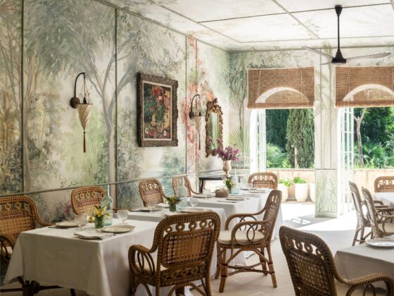 Sunlit dining area with white-clothed tables, wicker chairs, and a botanical mural wall.