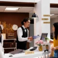 Receptionist in a white shirt and black vest hands a clipboard to a guest in a yellow top at the front desk in a hotel lobby