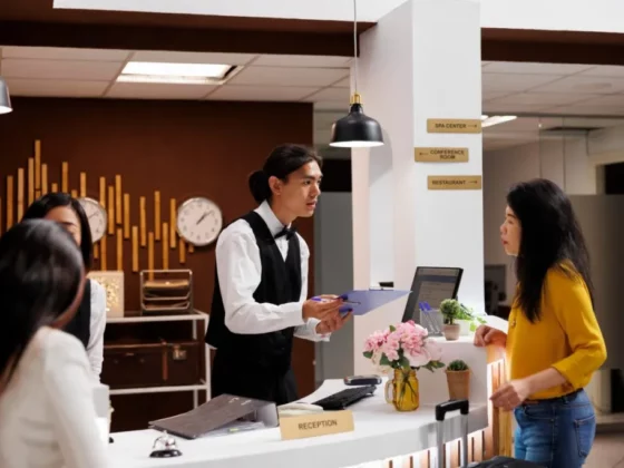 Receptionist in a white shirt and black vest hands a clipboard to a guest in a yellow top at the front desk in a hotel lobby