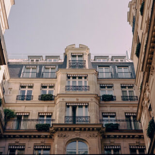Wide courtyard scene between ornate Parisian-style buildings with wrought-iron balconies and striped awnings, blue sky above.