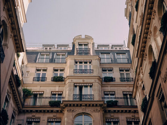 Wide courtyard scene between ornate Parisian-style buildings with wrought-iron balconies and striped awnings, blue sky above.