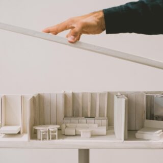 Hands place a long white strip over a white architectural scale model of a showroom with miniature furniture and cabinets.