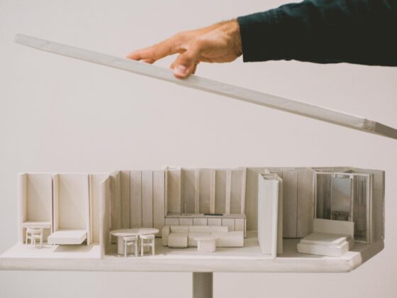 Hands place a long white strip over a white architectural scale model of a showroom with miniature furniture and cabinets.