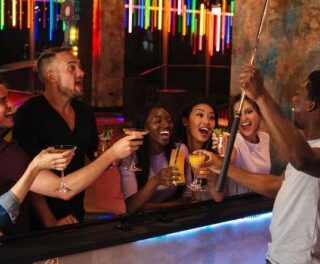 Group of friends at a bar taking a selfie with a selfie stick, raising drinks and laughing together.