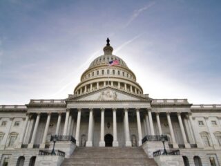 Front view of the U.S. Capitol with a flag on the dome, grand marble steps, and pressed-blue sky backdrop.