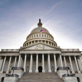 Front view of the U.S. Capitol with a flag on the dome, grand marble steps, and pressed-blue sky backdrop.