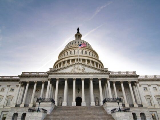 Front view of the U.S. Capitol with a flag on the dome, grand marble steps, and pressed-blue sky backdrop.