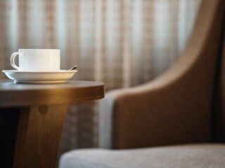 White coffee cup on a saucer with a spoon rests on a wooden table, blurred armchair in the background.
