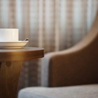 White coffee cup on a saucer with a spoon rests on a wooden table, blurred armchair in the background.
