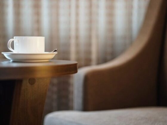 White coffee cup on a saucer with a spoon rests on a wooden table, blurred armchair in the background.