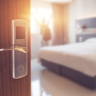 Close-up of a smart door lock on a wooden hotel room door, with a bright, celebratory figure in the blurred hotel room beyond.