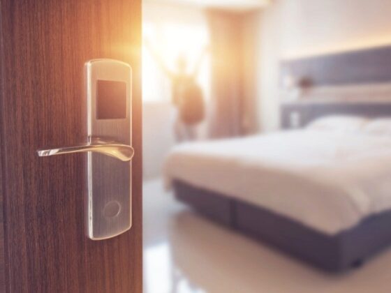 Close-up of a smart door lock on a wooden hotel room door, with a bright, celebratory figure in the blurred hotel room beyond.