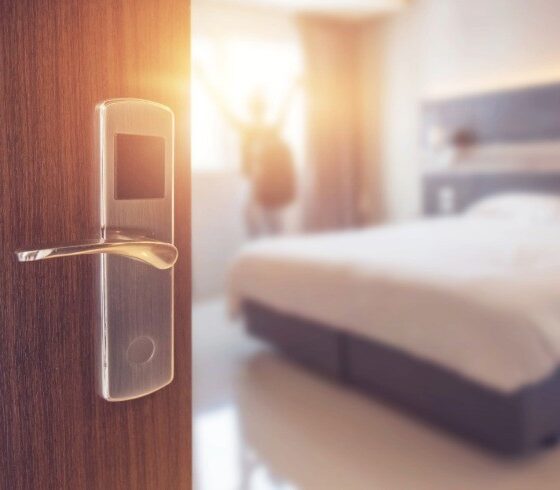 Close-up of a smart door lock on a wooden hotel room door, with a bright, celebratory figure in the blurred hotel room beyond.