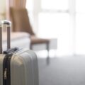 A silver hard-shell suitcase with extended handle standing on a carpeted floor beside a bed in a bright hotel room