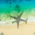 Aerial view of turquoise ocean meeting a sandy beach, with a large airplane shadow across the water near the shore.