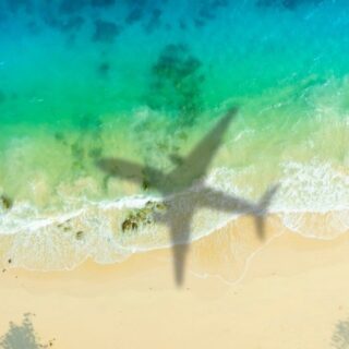 Aerial view of turquoise ocean meeting a sandy beach, with a large airplane shadow across the water near the shore.
