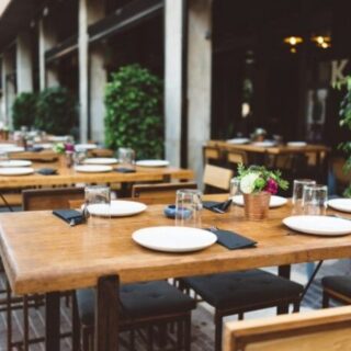Outdoor café with wooden tables set for dining, white plates, glasses, black napkins, and small flower centerpieces.