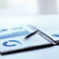 Printed business reports with blue bar and pie charts on a desk, a pen resting on the pages.