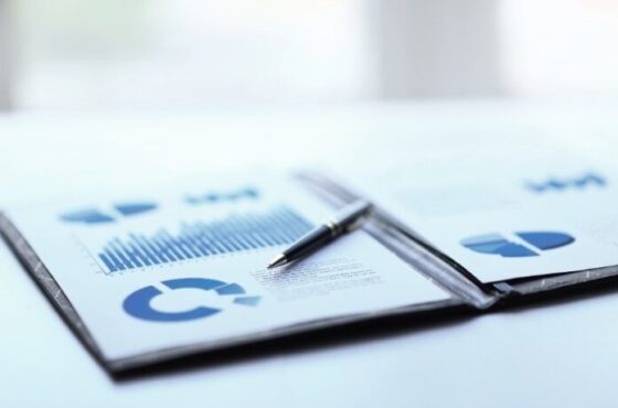 Printed business reports with blue bar and pie charts on a desk, a pen resting on the pages.