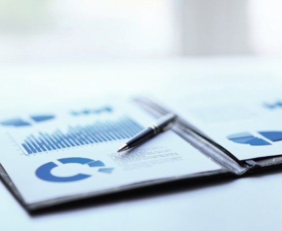 Printed business reports with blue bar and pie charts on a desk, a pen resting on the pages.