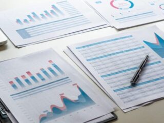 Several printed charts and spreadsheets with blue and red data visuals laid out on a desk, accompanied by a pen.