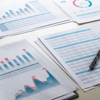 Several printed charts and spreadsheets with blue and red data visuals laid out on a desk, accompanied by a pen.