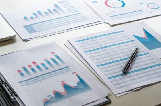 Several printed charts and spreadsheets with blue and red data visuals laid out on a desk, accompanied by a pen.
