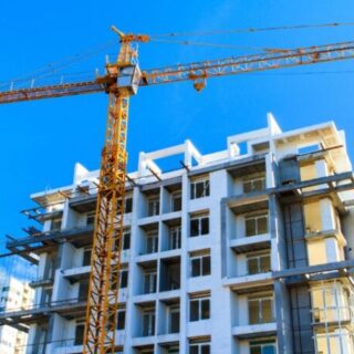 Tower crane looms over a partially completed white apartment building with exposed concrete floors and scaffolding, under a bright blue sky.