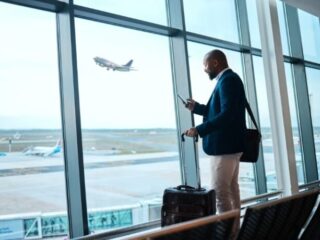 Businessman with rolling suitcase uses his phone at an airport terminal by large windows, as a plane takes off outside.