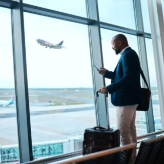 Businessman with rolling suitcase uses his phone at an airport terminal by large windows, as a plane takes off outside.