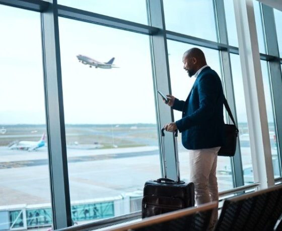 Businessman with rolling suitcase uses his phone at an airport terminal by large windows, as a plane takes off outside.