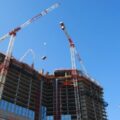 Two red-and-white construction cranes over a partially built high-rise with exposed concrete floors against a clear blue sky.